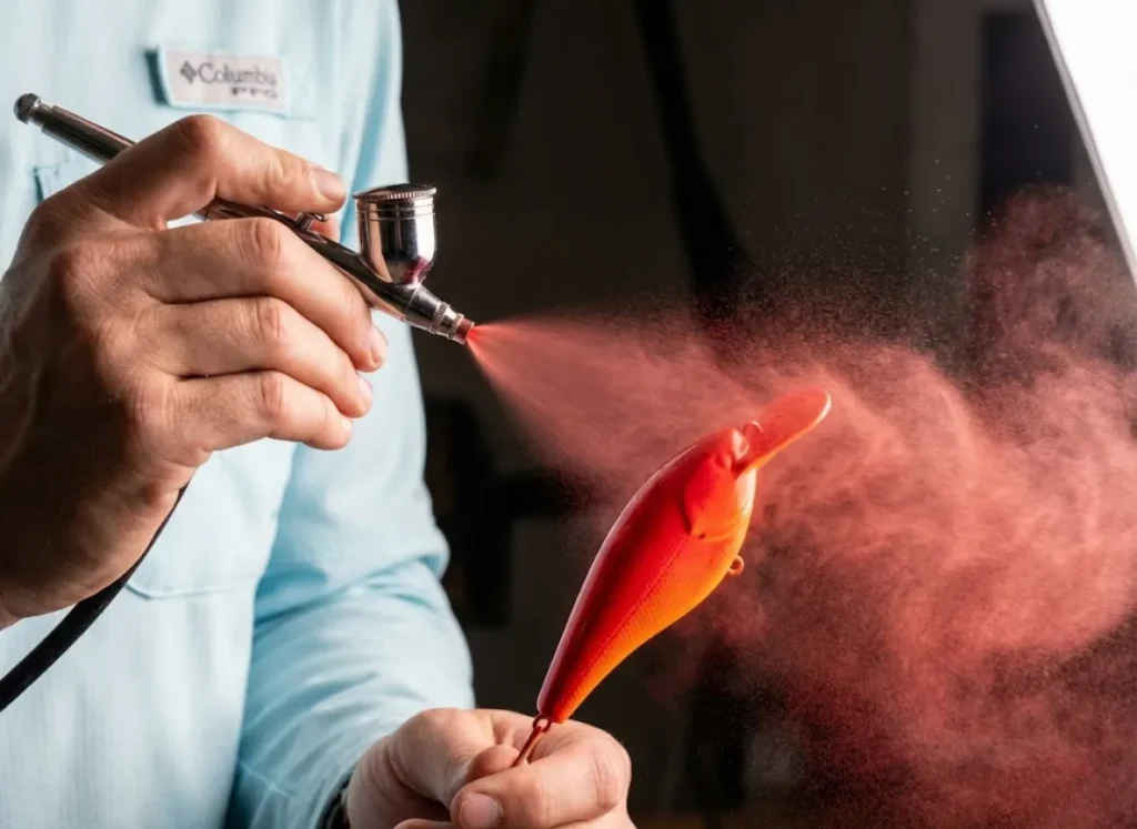 Action shot of an airbrush spraying red acrylic paint onto a fishing lure, capturing the fine mist and gradient application in high detail.