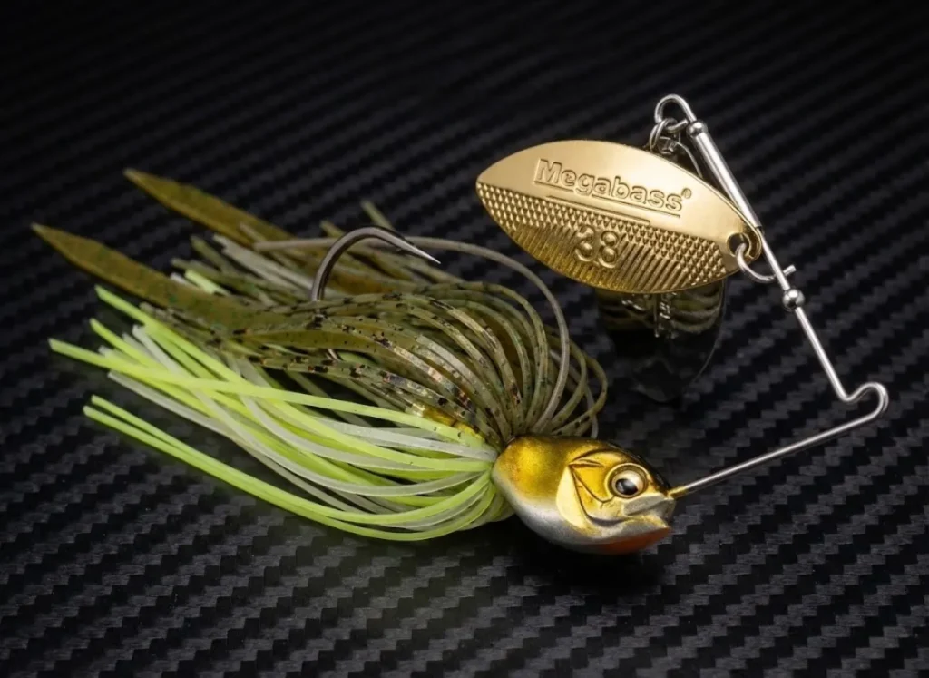 Macro product photography of a gold-bladed spinnerbait on a carbon fiber surface, highlighting the metallic texture and lure design.