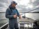 Control Your Boat Position in Heavy Wind A professional angler in a Simms jacket standing confidently on a boat deck in heavy wind and whitecaps, illuminated by dramatic storm sunlight.