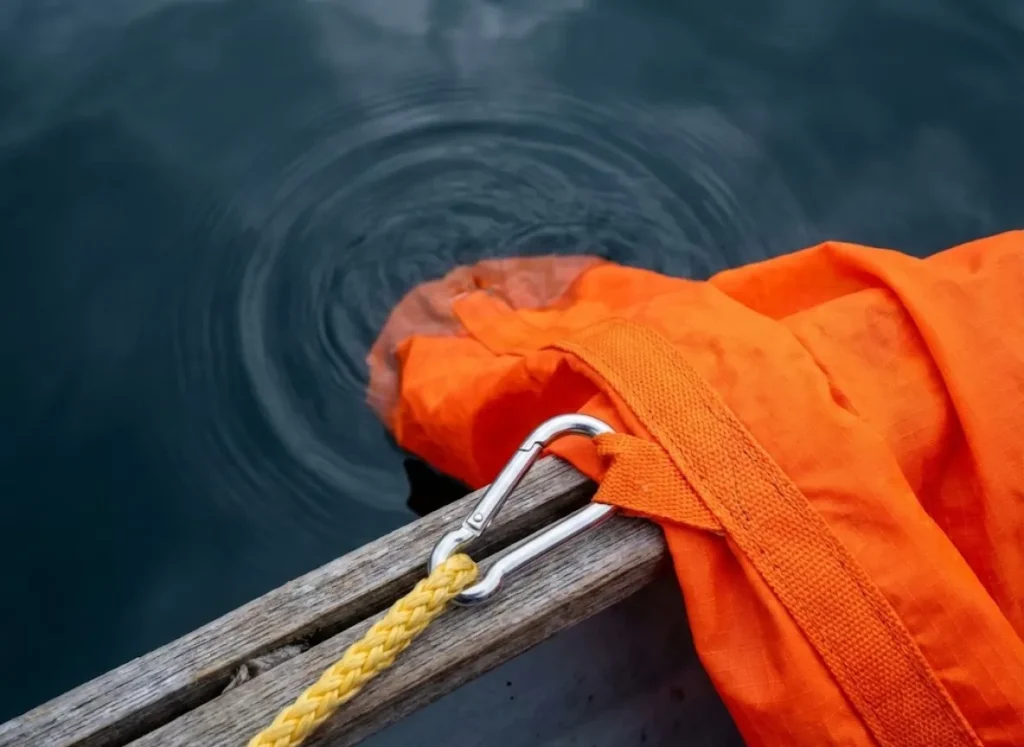 Top-down detail shot of a bright orange heavy-duty drift sock being lowered into dark water, highlighting the nylon fabric texture and stainless steel hardware.