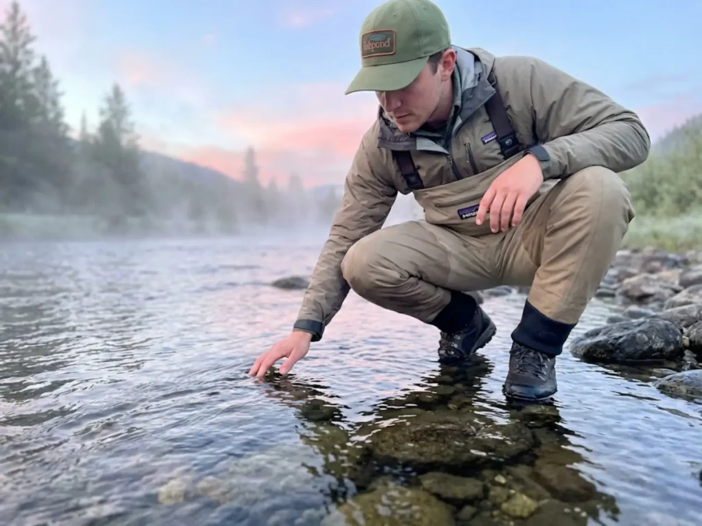 Angler studying fish behavior in clear stream water at dawn with morning mist