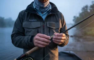 Use Hook Keepers to Prevent Transport Accidents A professional angler securing a lure to a hook keeper on a high-end fishing rod at dawn, wearing a Simms jacket, with a focus on gear safety and preparation.