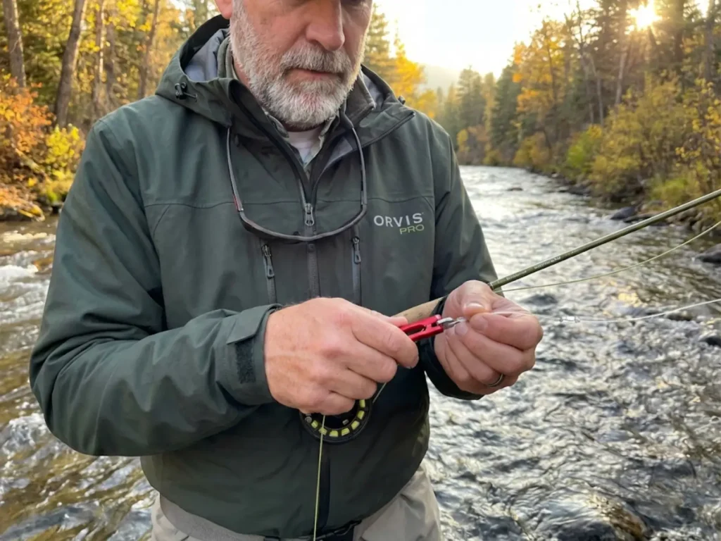 Fly angler using nail knot tool to attach leader to fly line on mountain river in autumn