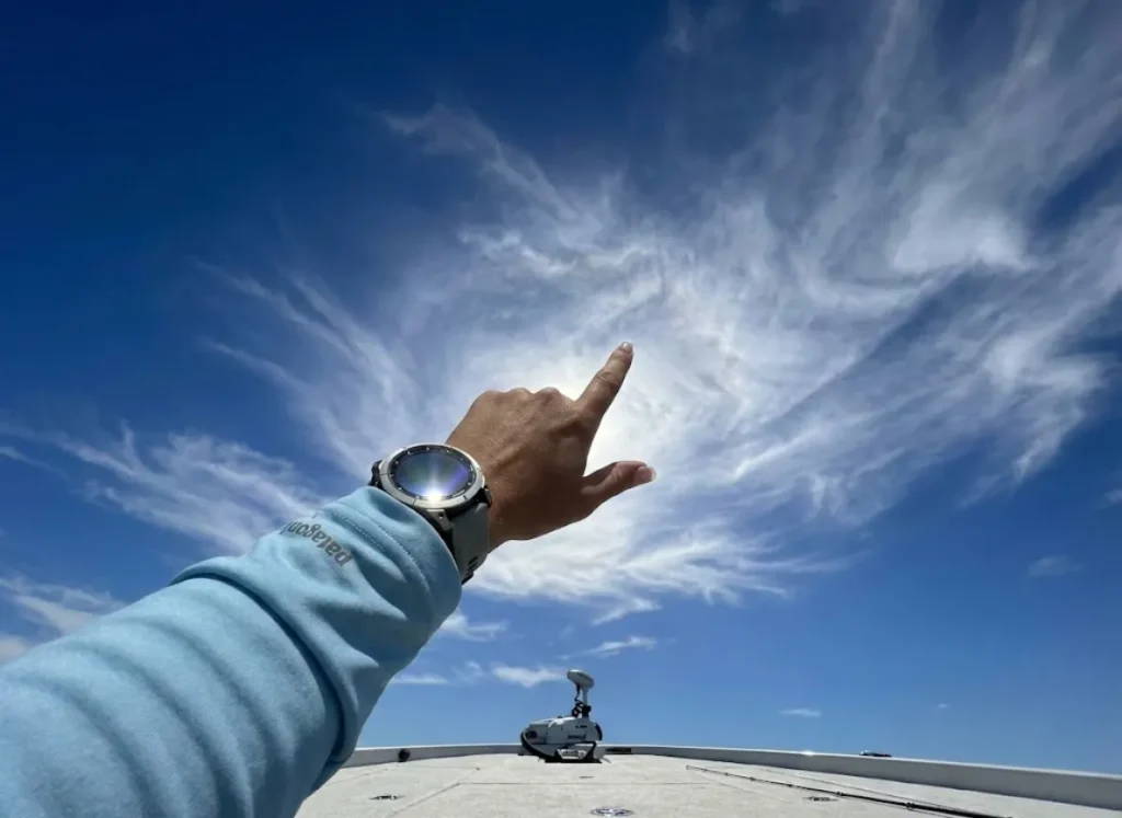 Angler pointing upward at cirrus clouds in a deep blue sky, wearing a Garmin watch and Patagonia sun hoodie, analyzing weather patterns.