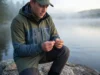 Prevent Lead Poisoning in Waterfowl and Wildlife A professional angler in Simms gear inspecting non-toxic tackle on a misty lake shoreline at dawn.