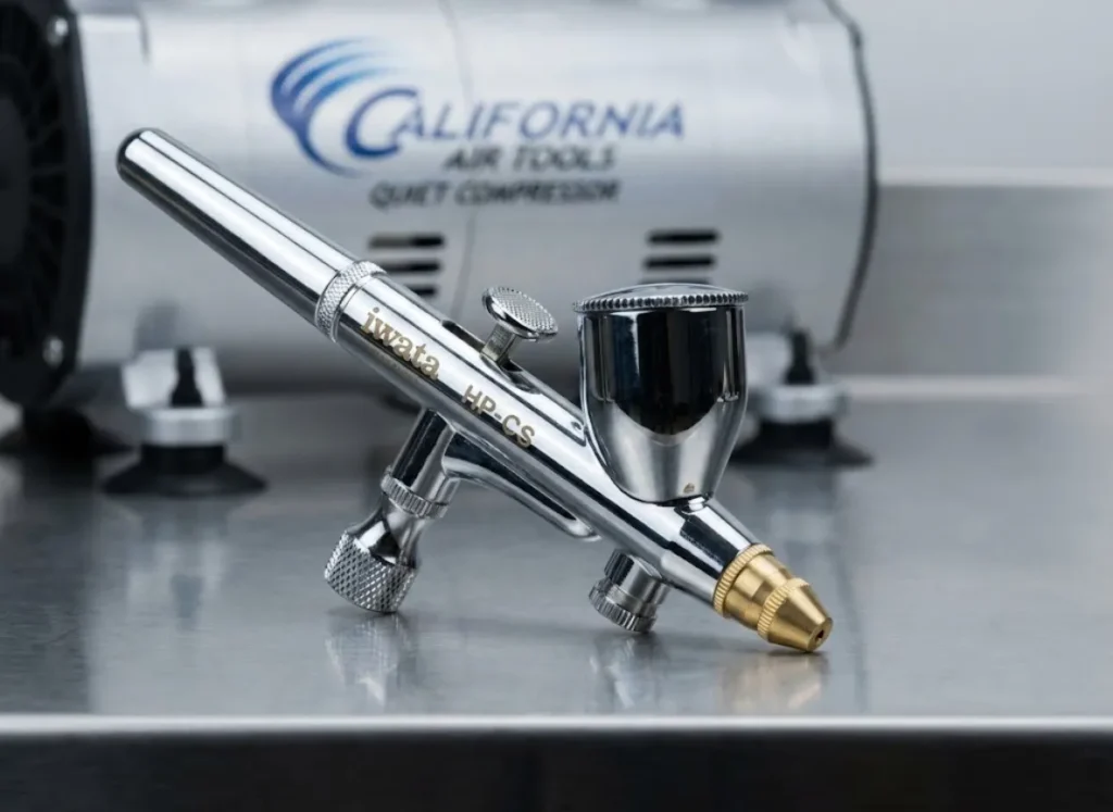 Macro photography of a chrome Iwata Eclipse HP-CS airbrush on a steel table, highlighting the knurled trigger and precision machining.