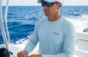 Recognize and Treat Heat Exhaustion on the Boat Professional angler at the helm of a boat under bright sun, wearing Simms protective gear and Costa sunglasses, representing maritime safety and preparedness.