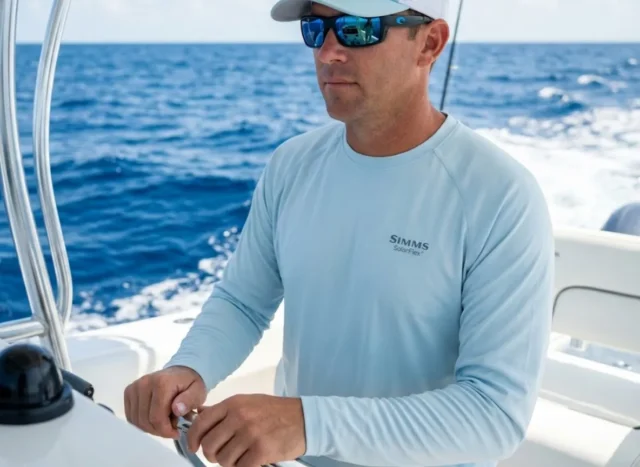 recognize-treat-heat-exhaustion-boat Professional angler at the helm of a boat under bright sun, wearing Simms protective gear and Costa sunglasses, representing maritime safety and preparedness.