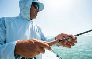 Spot Fish Before They See You A professional angler on the bow of a skiff in crystal clear shallow water, scanning the horizon with a fly rod in hand, wearing technical Simms fishing gear.