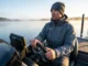 Track the Pre-Spawn Bass Transition Routes Professional angler in a Simms jacket scanning a misty lake at sunrise from a bass boat, representing the search for pre-spawn transition routes.
