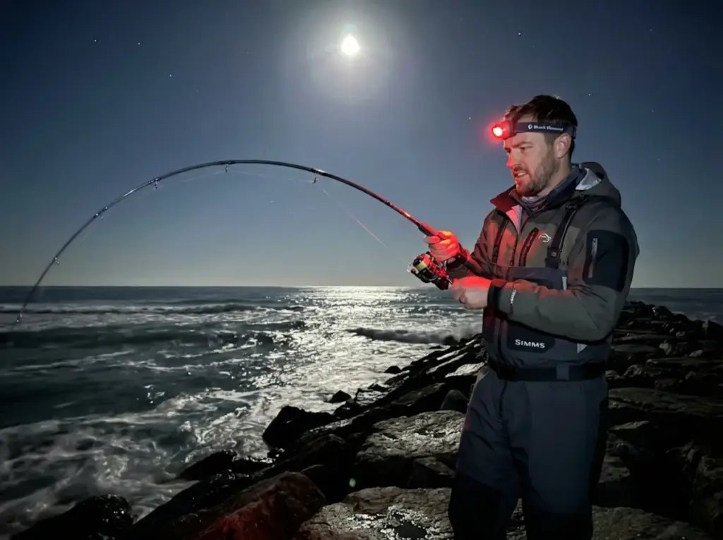 Angler fighting fish on coastal rocks under full moon using night fishing tactics