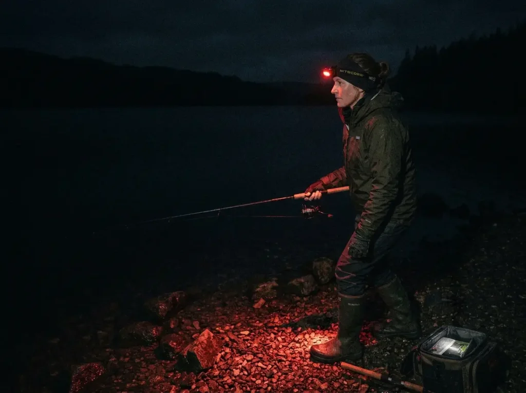 Night angler running dark with a red headlamp beam aimed at the bank, water left dark