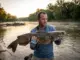 How I Finally Cracked the River Flathead Code Angler holding a large flathead catfish at dawn on a river bend with an Abu Garcia reel visible