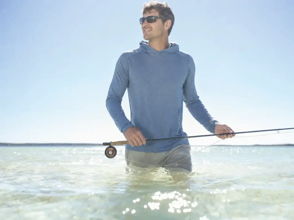 Angler wading shallow flats in a sun hoodie with UV glare reflecting off the water surface