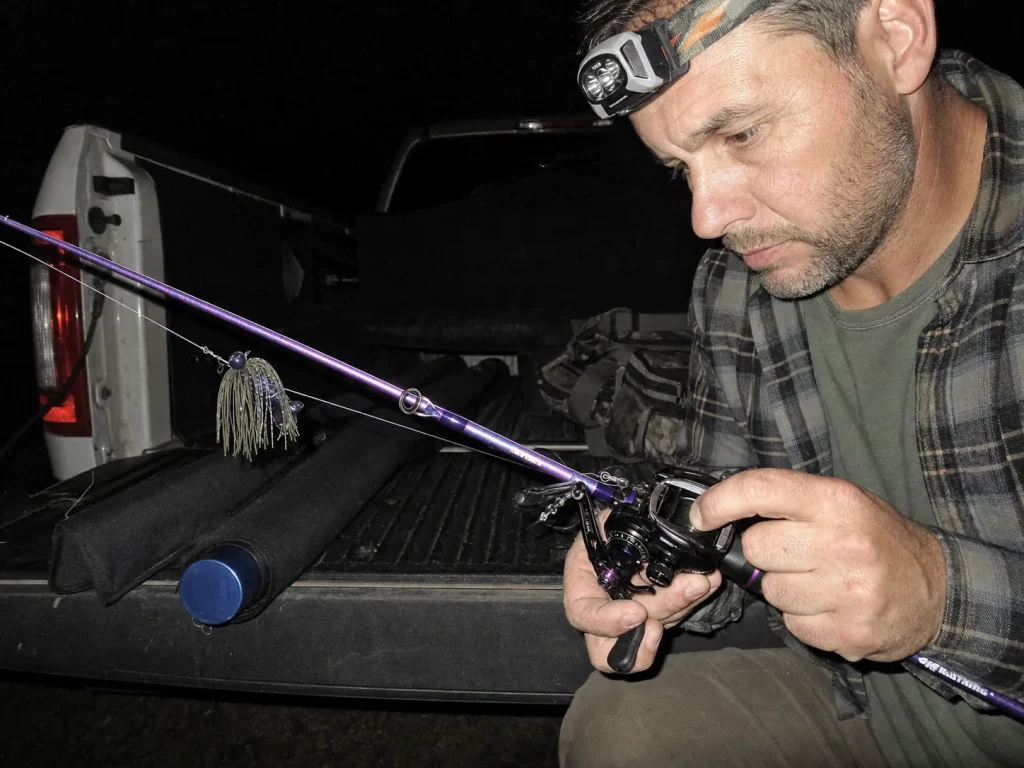 Angler adjusting baitcaster spool tension by flashlight at a truck tailgate before dawn