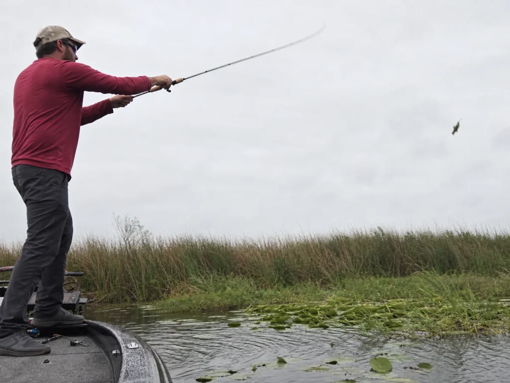 Angler working a jig along a grassy shoreline edge in stained water from a boat