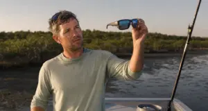 Fishing Sunglasses Lens Color Types Explained Angler holding polarized fishing sunglasses up to the light to check lens color on a skiff deck