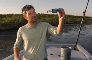 Fishing Sunglasses Lens Color Types Explained Angler holding polarized fishing sunglasses up to the light to check lens color on a skiff deck