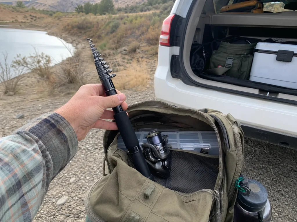 A collapsed telescoping fishing rod tucked into a daypack side pocket beside a truck door on a road trip