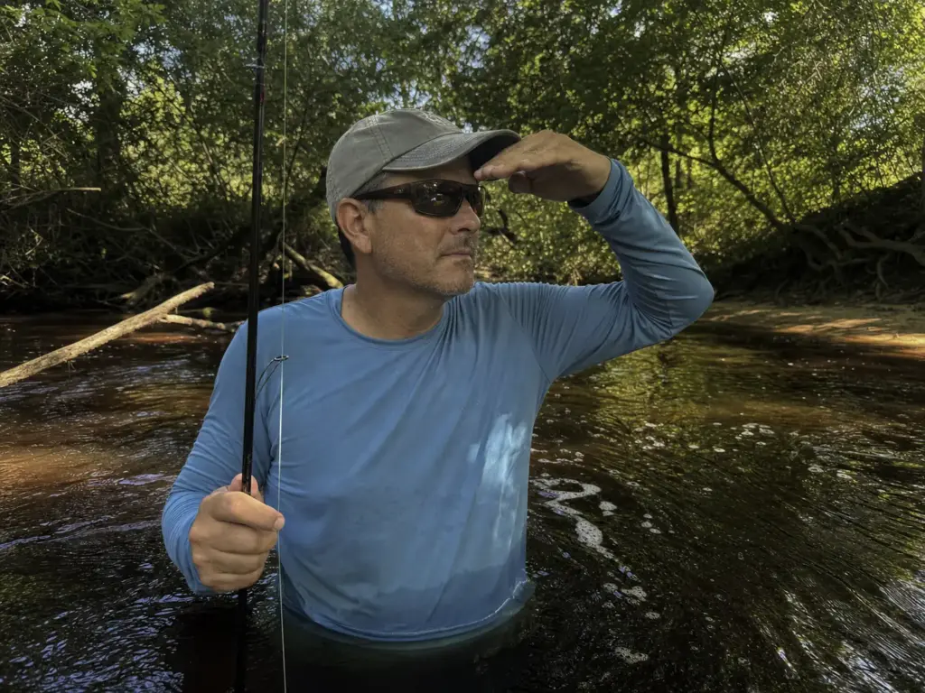 Wading angler in copper polarized lenses reading tannin-stained backwater under tree cover