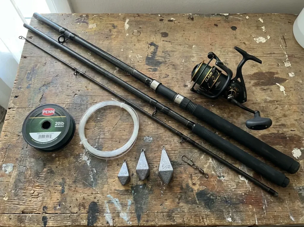 Pendulum cast gear flat-lay: 13-foot surf rod, spinning reel, braid mainline, shock leader, and pyramid sinkers laid out on a wooden bench