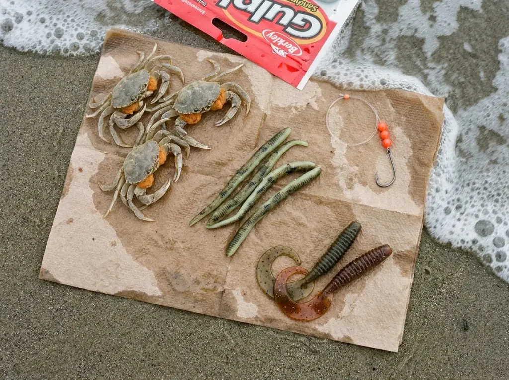 Selection of surf perch baits including sand crabs, Berkley Gulp sandworms, and curlytail grubs on wet sand