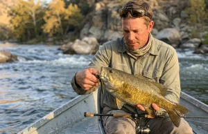 7 Tube Jig Retrieves That Crack Smallmouth Angler holding smallmouth bass caught on tube jig over rocky river, green pumpkin tube visible