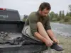 Best Fishing Socks for Sweaty Feet in Waders Angler pulling a merino wool fishing sock onto his foot at a river access before putting on his waders
