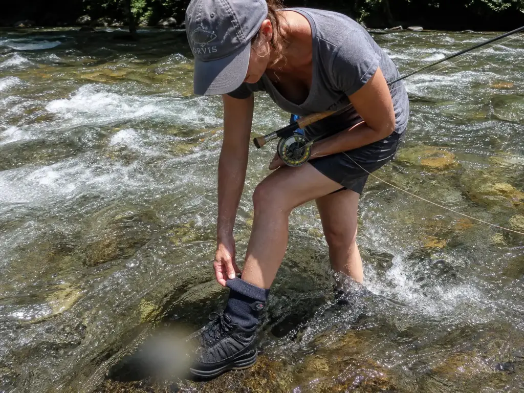 Wet wading angler in a rocky river adjusting the cuff of a neoprene wet wading sock above her boot
