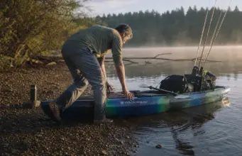 Fishing Kayak Buying Guide What Actually Matters Angler pushing a rigged fishing kayak off a gravel bank at first light