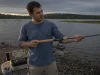 How to Choose a Fishing Rod Without Wasting Money Angler holding a spinning rod at a lake shore while deciding how to choose a fishing rod
