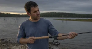 How to Choose a Fishing Rod Without Wasting Money Angler holding a spinning rod at a lake shore while deciding how to choose a fishing rod