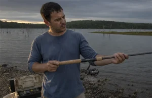 How to Choose a Fishing Rod Without Wasting Money Angler holding a spinning rod at a lake shore while deciding how to choose a fishing rod