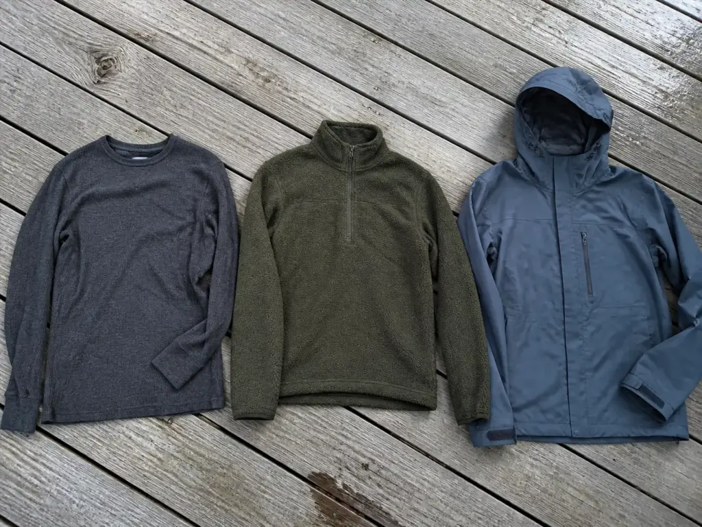 Three cold-weather fishing layers laid out flat on a wooden dock — merino base layer, fleece mid layer and waterproof shell