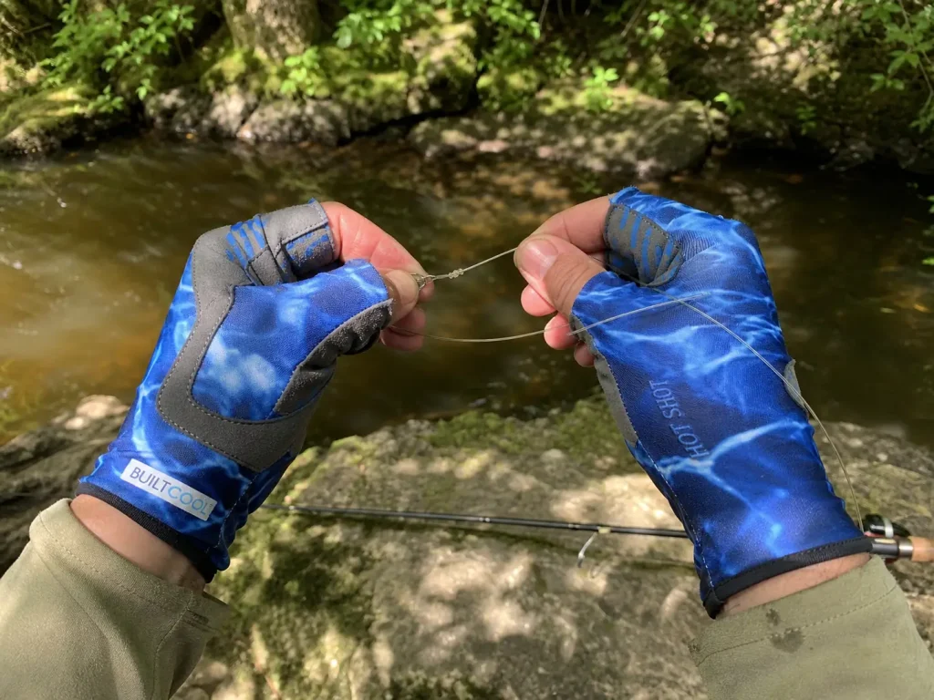 Fingerless UPF sun gloves on an angler's hands tying a knot on a riverbank in dappled light